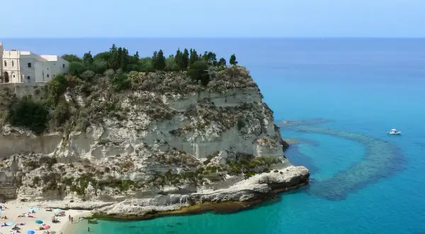 Scogliera bianca con un santuario in cima, bagnata da un mare cristallino in Calabria. Spiaggia affollata e barca all'orizzonte.