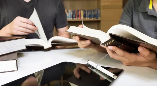 Estudiantes consultan libros y tablet en biblioteca, preparándose para las competencias específicas LOMLOE Andalucía.