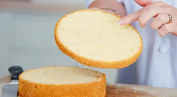 Perfekter tortenboden rezept: Eine Hand hebt eine goldbraune Kuchenschicht über eine zweite, die auf einem Holzbrett liegt.