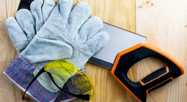 Guantes anticorte niveles, gafas de seguridad y sierra de mano sobre madera.