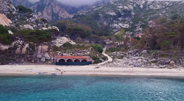 Spiaggia selvaggia con rocce e vegetazione, un edificio storico e montagne nebbiose sullo sfondo. Qui si trova l'isola di Montecristo.