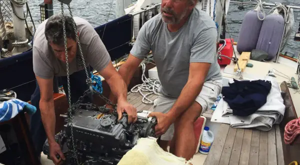 Due uomini lavorano alla manutenzione del motore di una barca a vela, sollevandolo con una catena.
