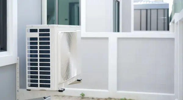 Unité extérieure d'une pompe à chaleur appartement, fixée sur un mur blanc. L'appareil est blanc avec des grilles bleues et un ventilateur visible.