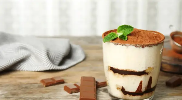 Szybki deser z niczego: kremowe tiramisu w szklance, posypane kakao i udekorowane miętą. Obok kawałki czekolady.
