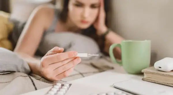 Chora dziewczyna z termometrem w ręku, leżąca w łóżku. Obok leki i kubek herbaty. L4 online 49 zł.