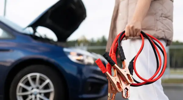 Osoba trzyma kable rozruchowe przy samochodzie z otwartą maską. Dowiedz się, jak podłączyć kable rozruchowe, by uruchomić auto.