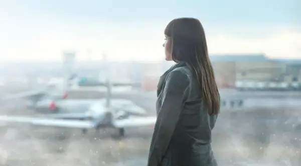 Dziewczyna w szarym płaszczu patrzy przez okno na lotnisku, gdzie w oddali widać samolot Boeing 737-8.