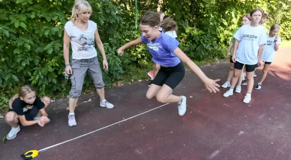 Dziewczynka skacze przez linkę podczas lekcji wychowania fizycznego w szkole. Nauczycielka i inne dzieci obserwują.