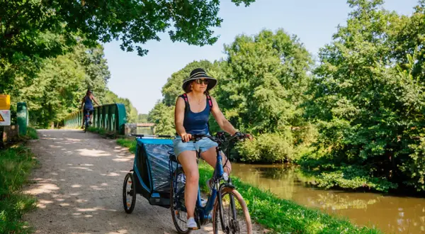 Mujer pedalea por las vías verdes de Francia con un remolque para niños, disfrutando del paisaje junto al canal.