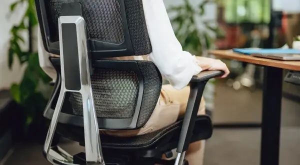 Person in bequemer Sitzposition am Schreibtisch, die Armlehne des Bürostuhls nutzend.
