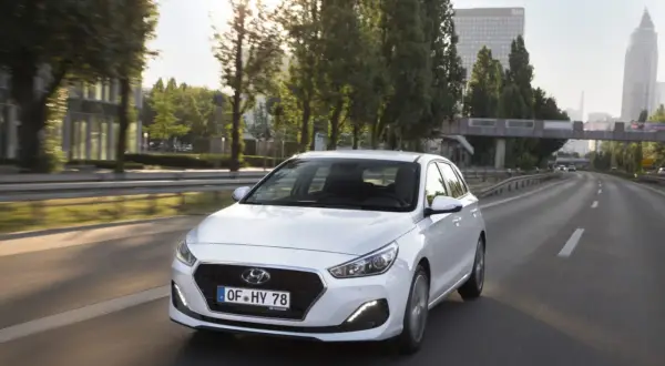 Biały Hyundai i30 1.4 benzyna na drodze. Kierowca w okularach przeciwsłonecznych. Opinie o tym modelu są pozytywne.