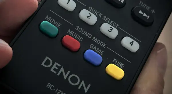 Dłoń trzyma pilot Denon. Widoczne przyciski: Movie, Music, Game, a także żółty przycisk PURE.