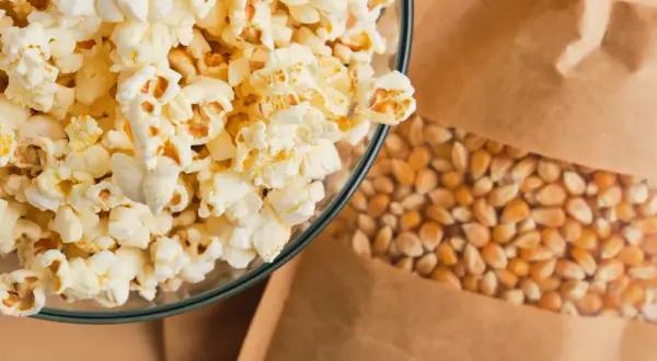 Miseczka pełna puszystego popcornu obok ziaren kukurydzy w papierowej torbie. Czy popcorn jest lekkostrawny? Zależy od dodatków!