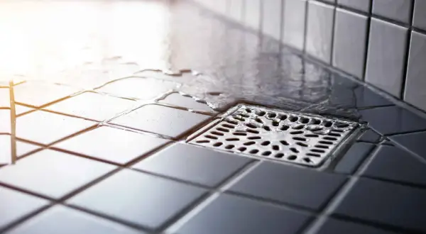 Acqua stagnante vicino allo scarico doccia puzza. Le piastrelle scure e bagnate creano un'atmosfera poco invitante.
