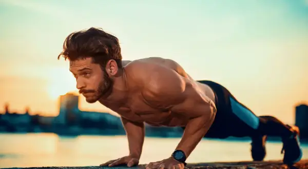 Mężczyzna wykonuje pompki na biceps przy zachodzie słońca nad wodą.
