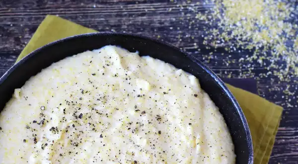 Krämig italiensk polenta i en gjutjärnspanna, strödd med svartpeppar. Majsgryn syns på den mörka träytan.