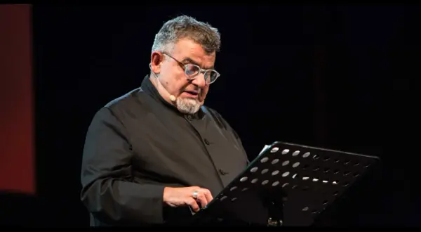 Giancarlo Cauteruccio, con occhiali e barba, legge da un leggio su un palco buio.