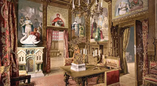 Prunkvoller Salon im Schloss Neuschwanstein innen. Wandmalereien mit Sagenmotiven, ein Kamin mit Schwan, prunkvolle Möbel und ein Kronleuchter schaffen königliches Ambiente.