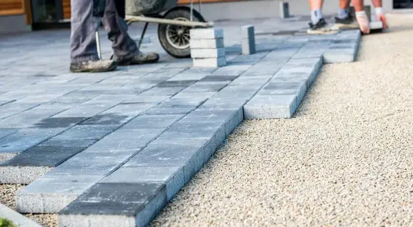 Układanie kostki brukowej, tworząc ścieżkę, która jest co zamiast krawężnika. W tle widać pracujących ludzi i taczkę.