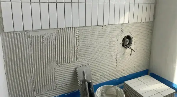 Jak położyć płytki w łazience: klej na ścianie, kielnia, wiaderko z zaprawą i stos białych płytek.