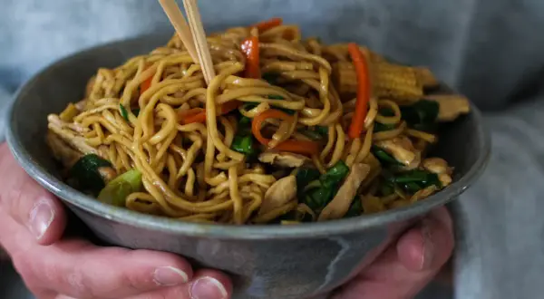 En skål med lo mein-nudlar, kyckling och grönsaker, lyfts med ätpinnar.