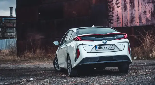 Biały Prius 4 z napędem hybrydowym stoi na tle industrialnego krajobrazu.