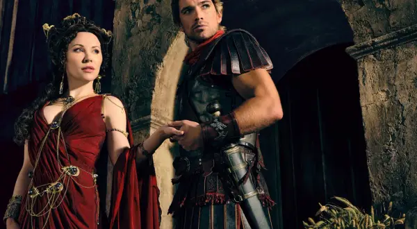 Die Besetzung von Spartacus: Ashur und Lucretia, in prachtvollen Gewändern und Rüstungen, halten Händchen vor einer antiken Kulisse.