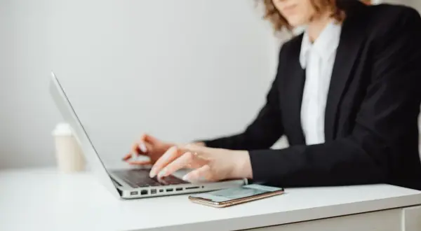 Kobieta w okularach i czarnej marynarce pracuje na laptopie. Obok leży telefon. Może szuka informacji, co to jest upadłość konsumencka.