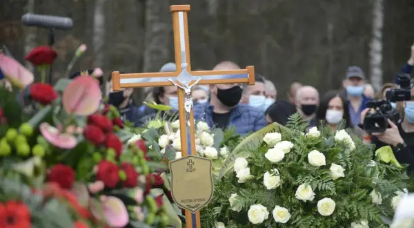 Drewniany krzyż z Jezusem, otoczony wieńcami, symbolizuje pożegnanie. Po pogrzebie tabliczka z napisem "Spoczywaj w pokoju" pozostaje jako pamiątka.