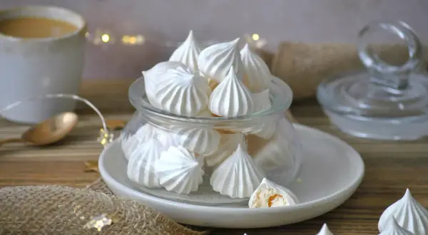Leckeres Meringen Rezept: zarte Baiser-Häufchen in einem Glas, daneben eine Tasse Kaffee und Lichterkette.