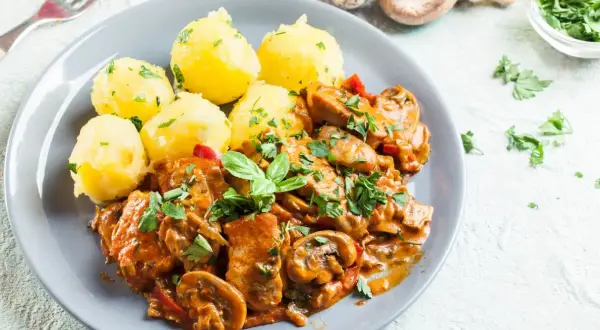 Pyszny gulasz z kurczaka z pieczarkami i papryką, podany z ziemniakami posypanymi natką pietruszki.