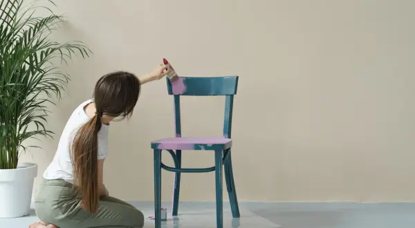 Donna inginoxida una sedia blu con vernice lucida, applicando un colore rosa pastello per opacizzare la finitura.