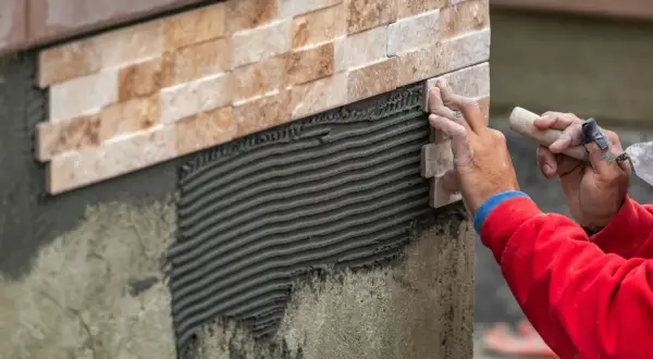 Close-up of a worker's hands carefully performing wall tile installation, applying a new stone tile to a textured adhesive layer.