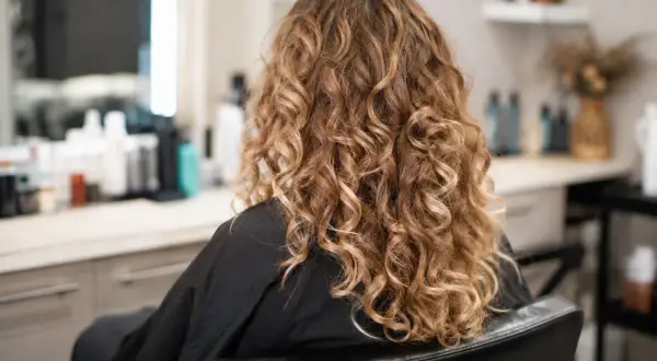 Kobieta w czarnym szlafroku, z pięknymi, złotymi loki trwała. W tle widać salon fryzjerski.