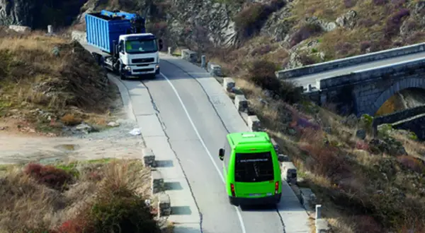 Camión blanco con contenedor azul subiendo una pendiente, seguido por un autobús verde. Se debate quién tiene prioridad en una pendiente.