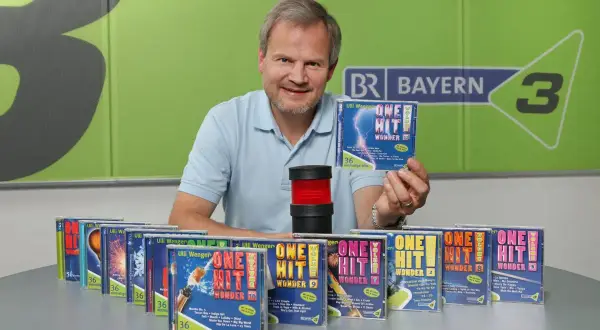 Mann präsentiert CDs mit dem Titel "One Hit Wonder". Viele weitere CDs dieser Reihe sind auf dem Tisch aufgereiht.