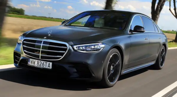 Elegancki, ciemnoszary Mercedes S580e sunie po drodze wśród zielonych pól i drzew.