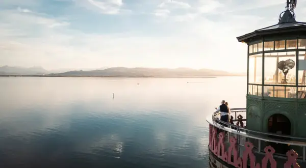 Ein Paar genießt den Sonnenuntergang am Leuchtturm Lindau. Der Bodensee glänzt, Berge im Hintergrund.
