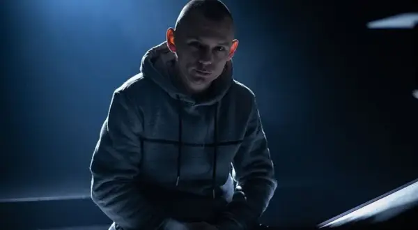 Mężczyzna w szarej bluzie z kapturem, oświetlony od góry, na ciemnym tle.