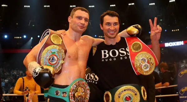 Two boxers, one with multiple championship belts, stand together. The boxer on the left, Vitali Klitschko, shows his impressive height and championship status.