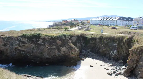 Acantilados rocosos y una pequeña cala de arena en la playa de San Bartolo Barreiros, con edificios residenciales al fondo.