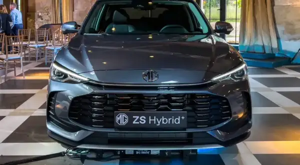 Szary MG ZS Hybrid+ prezentuje się elegancko na tle wnętrza z zasłonami i krzesłami.