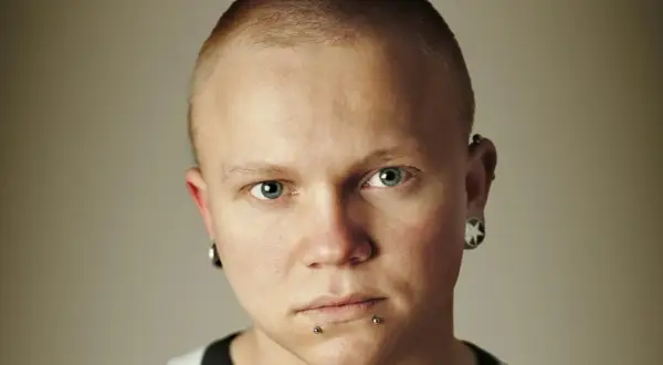 Mężczyzna z niebieskimi oczami i kolczykiem w nosie. Ma krótko ostrzyżone włosy i kolczyki w uszach.