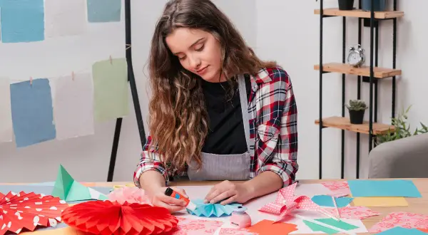 Chica creando manualidades para niños de 12 a 15 años. Pliegues de papel, abanicos y adornos coloridos sobre la mesa.