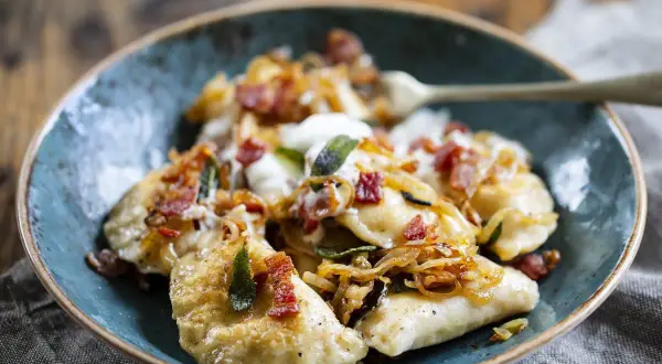 Pyszne pierogi z serem, okraszone skwarkami i cebulką, podane w niebieskiej misce.