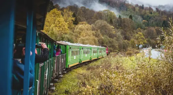 Zielony pociąg PKP Ustrzyki Dolne w jesiennym krajobrazie. Turyści podziwiają widoki, sprawdzając rozkład jazdy.