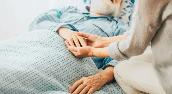 Ein älterer Mann mit Bart und Nasenbrille liegt im Bett. Eine Person hält seine Hand. Die Szene vermittelt die Endphase, wie man an Prostatakrebs stirbt.