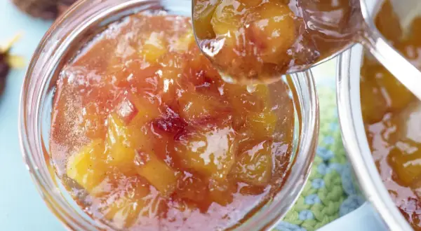 Ein Löffel voller glänzender Nektarinen Marmelade wird über ein Glas voller Fruchtstücke geschöpft.