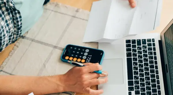Osoba analizuje dokumenty, korzystając z kalkulatora w telefonie i laptopa, by obliczyć maksymalne oprocentowanie kredytu.
