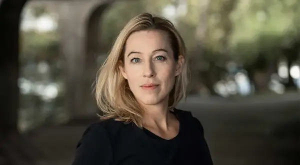 Anna Careborg, en kvinna med blont hår och blå ögon, tittar rakt fram. Bakgrunden är suddig med grönska och en valvformad struktur.